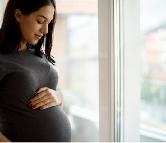 Schwangere steht am Fenster und berührt ihren Bauch