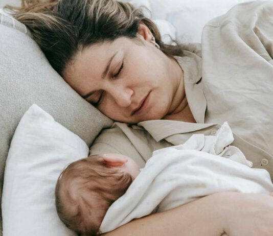 Erschöpfte Mama liegt mit schlafendem Baby im Arm.