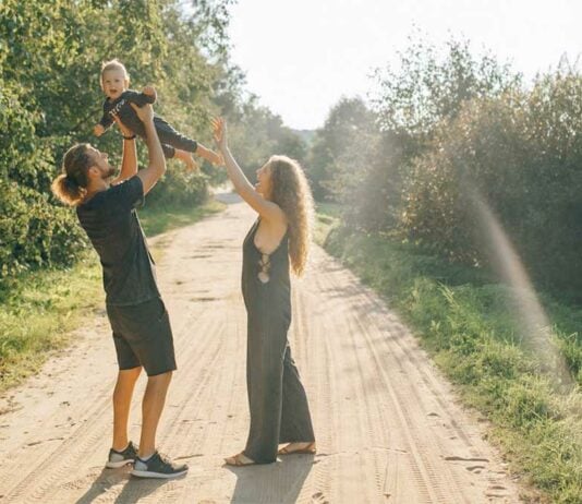Mama und Papa stehen auf sonnigem Feldweg und werfen lachendes Kind in die Luft.