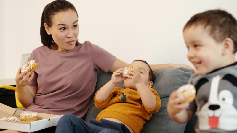 Mutter sitzt mit ihren Kindern auf dem Sofa und alle essen Spritzkuchen.