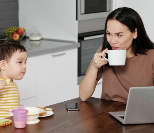 Mutter sitzt mit ihrem Kind am Esstisch und trinkt Kaffe während das Kind frühstückt. Dabei hat die Mutter ihren Laptop vor sich.