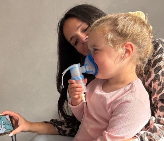 Kleines Mädchen inhaliert nach einer Infektion mit RSV und einer Bronchitis mit dem Inhalationsgerät von Pari. Sie sitzt dabei auf dem Schoß von ihrer Mutter.