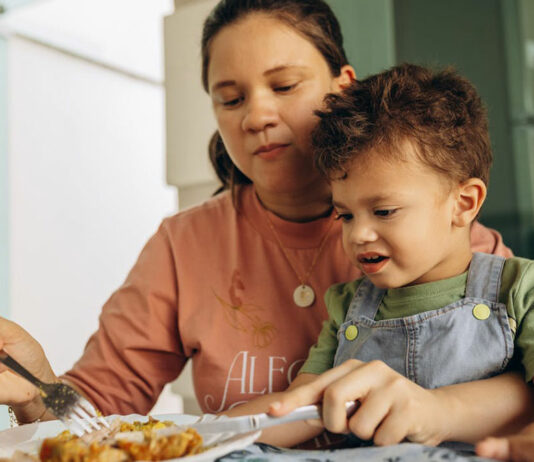 Mama versucht skeptisch schauendes Kind zu füttern.