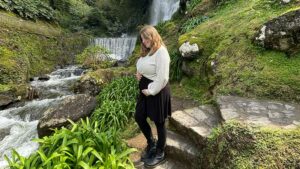 Laura steht vor einem Wasserfall und hält ihren schwangeren Bauch. 