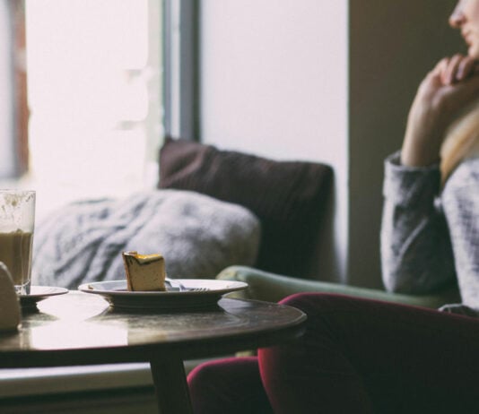 Frau sitz auf einem Sessel und schaut nachdenklich aus dem Fenster. Vor ihr auf dem Tisch stehen Kuchen und Kaffee.