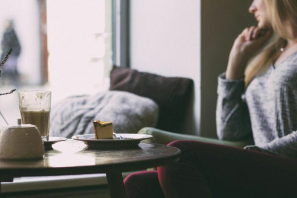 Frau sitz auf einem Sessel und schaut nachdenklich aus dem Fenster. Vor ihr auf dem Tisch stehen Kuchen und Kaffee.