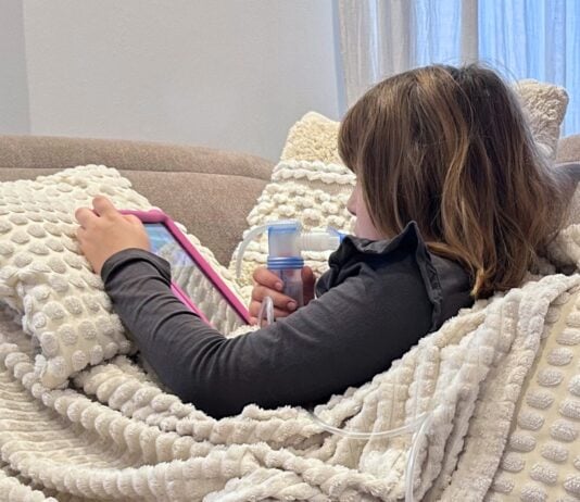 Mädchen inhaliert auf der Couch mit dem Inhalaitonsgerät PARI COMPACT2 und schaut dabei eine Serie.