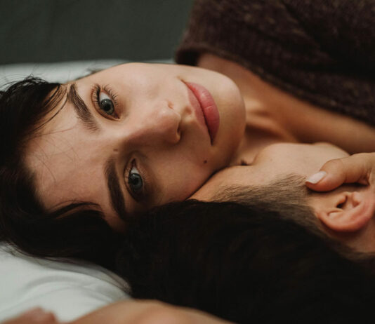 Paar liegt im Bett. Frau schaut verträumt in die Ferne und berührt ihn am Hals.