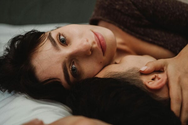 Paar liegt im Bett. Frau schaut verträumt in die Ferne und berührt ihn am Hals.