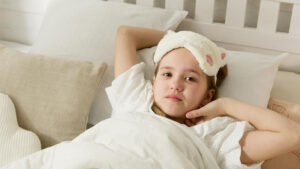 Mädchen liegt wach mit Schlafmaske auf der Stirn in ihrem Bett.