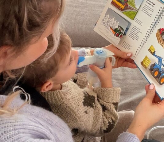 Kind sitzt auf dem Schoß von der Mama beim Inhalieren, während sie ein Buch vorliest.