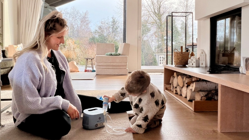 Mama und Kind sitzen im Wohnzimmer auf dem Boden während das Kind das Inhalationsgerät einschaltet.